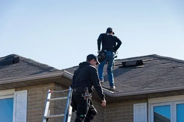 Two roofers on a rooftop, one on a ladder, working on a gray shingled roof under a clear blue sky.