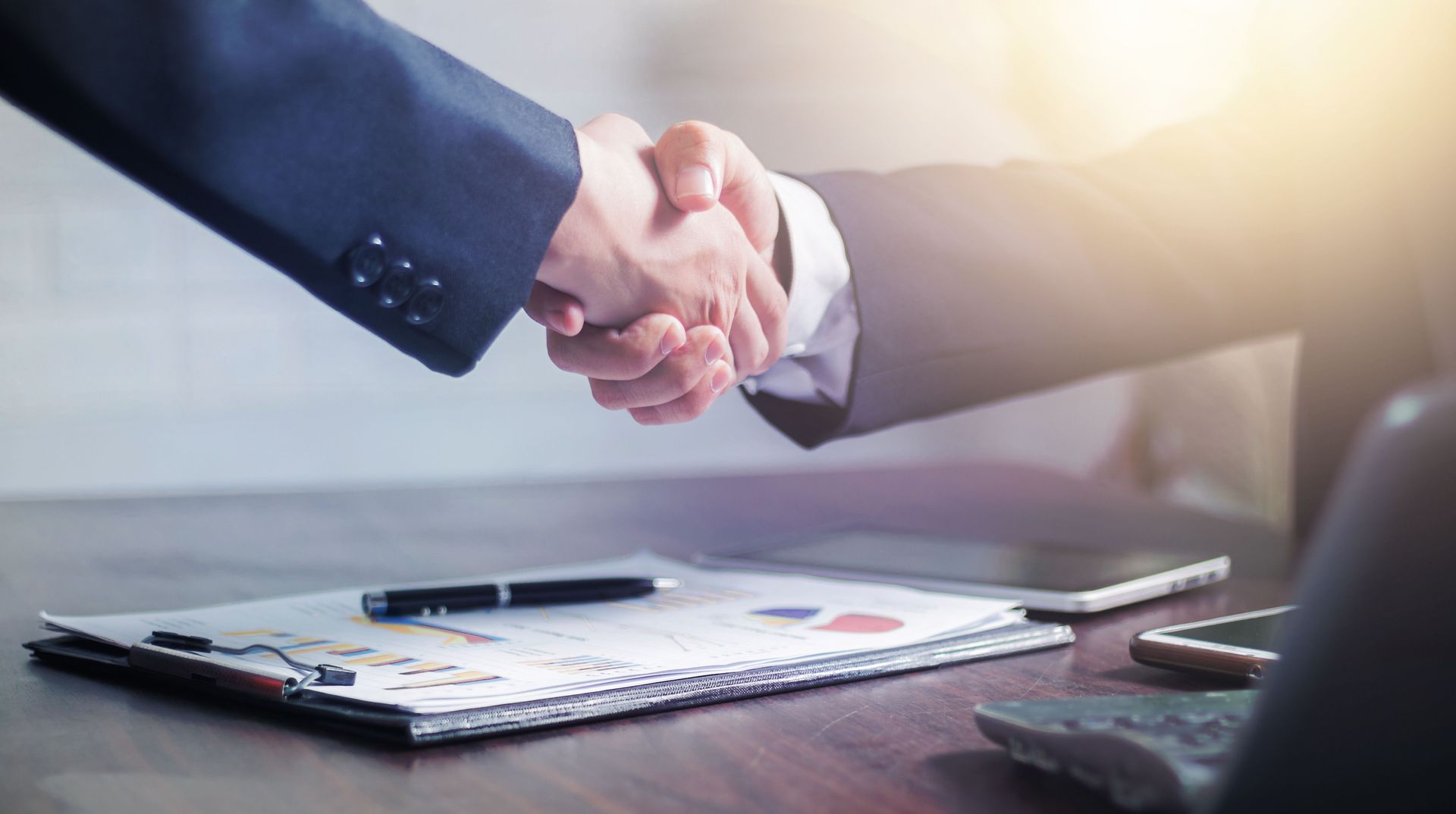 Businesspeople shaking hands over a desk with charts and a pen under warm office light