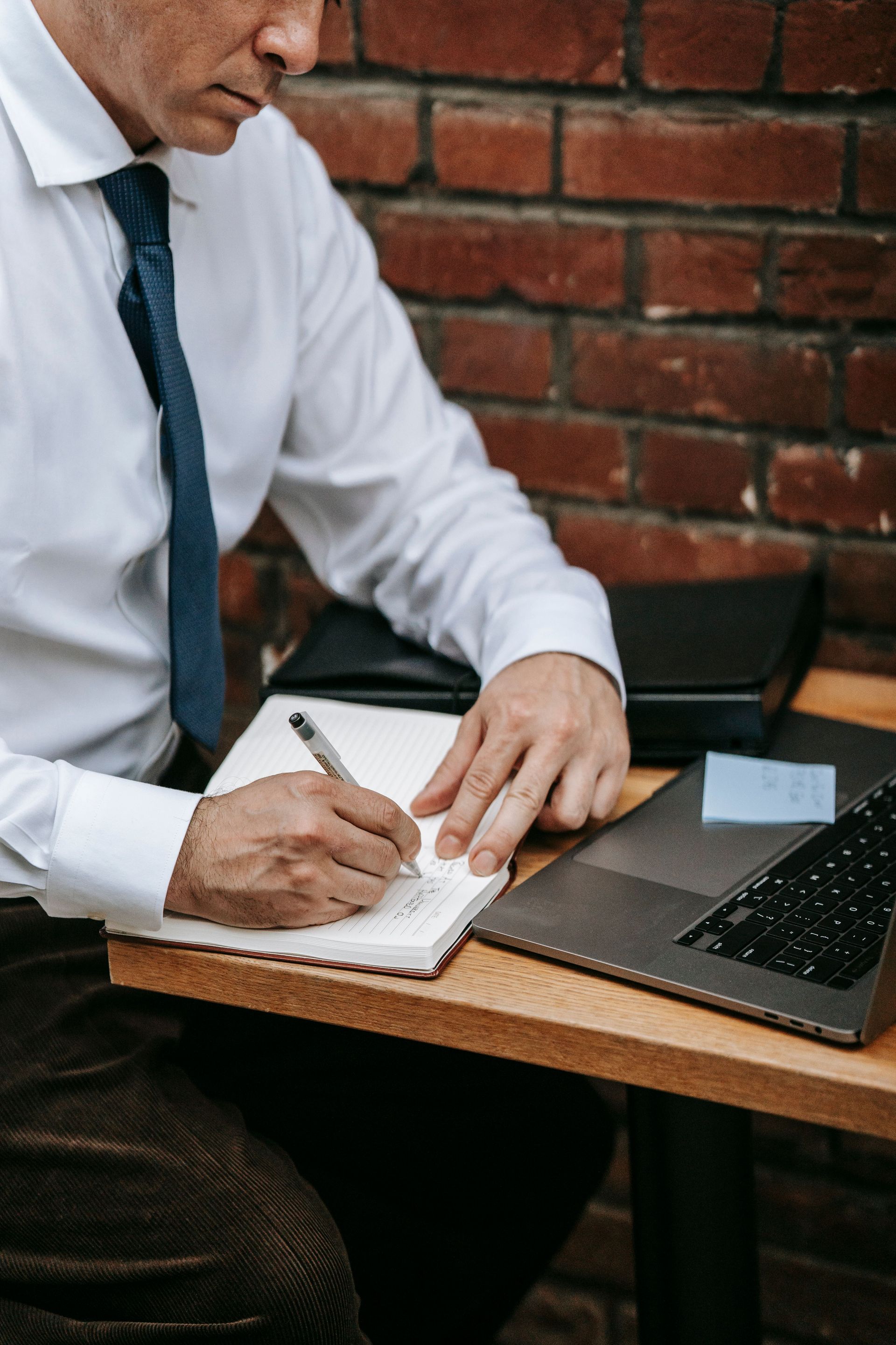 Person writing in a notebook beside a laptop at a wooden table, with a brick wall background.