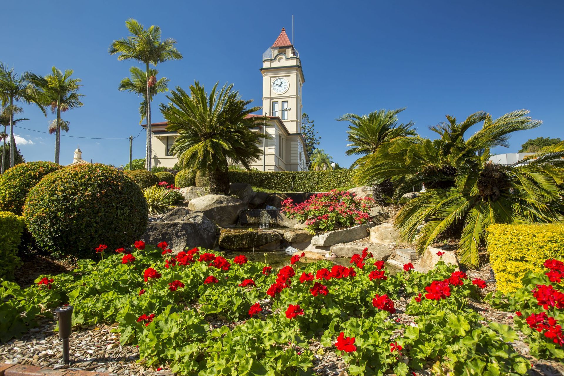 A Clock Tower Surrounded by Red Flowers and Lush Greenery — Country To Coast Building Contractors In Gympie, QLD
