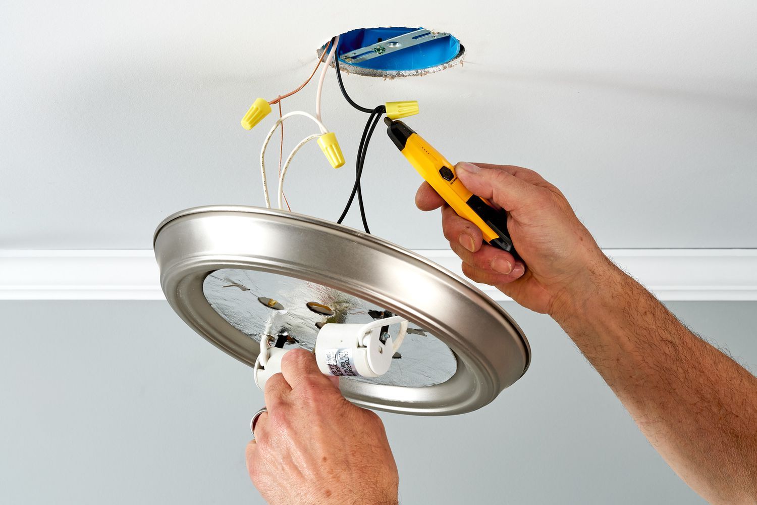 A person installing a ceiling light fixture with wires and tools in hand.