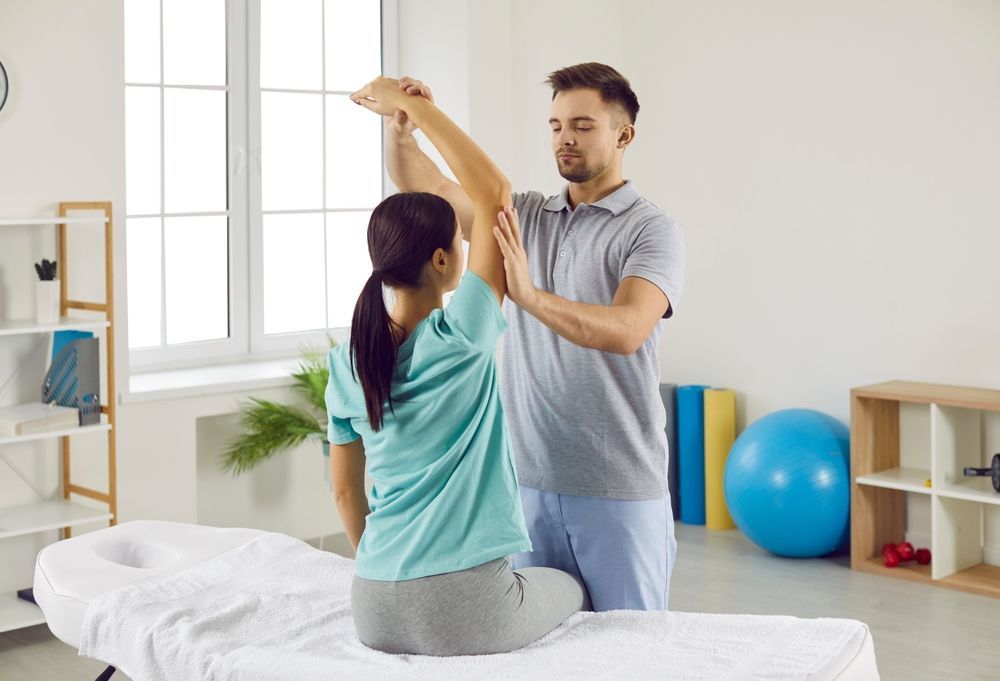 Therapist assisting a patient with arm movement in a bright therapy room.
