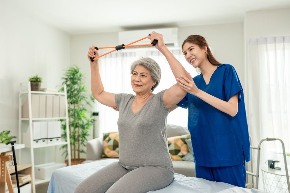 Woman using resistance band, supervised by a healthcare worker. Indoors, they are smiling.