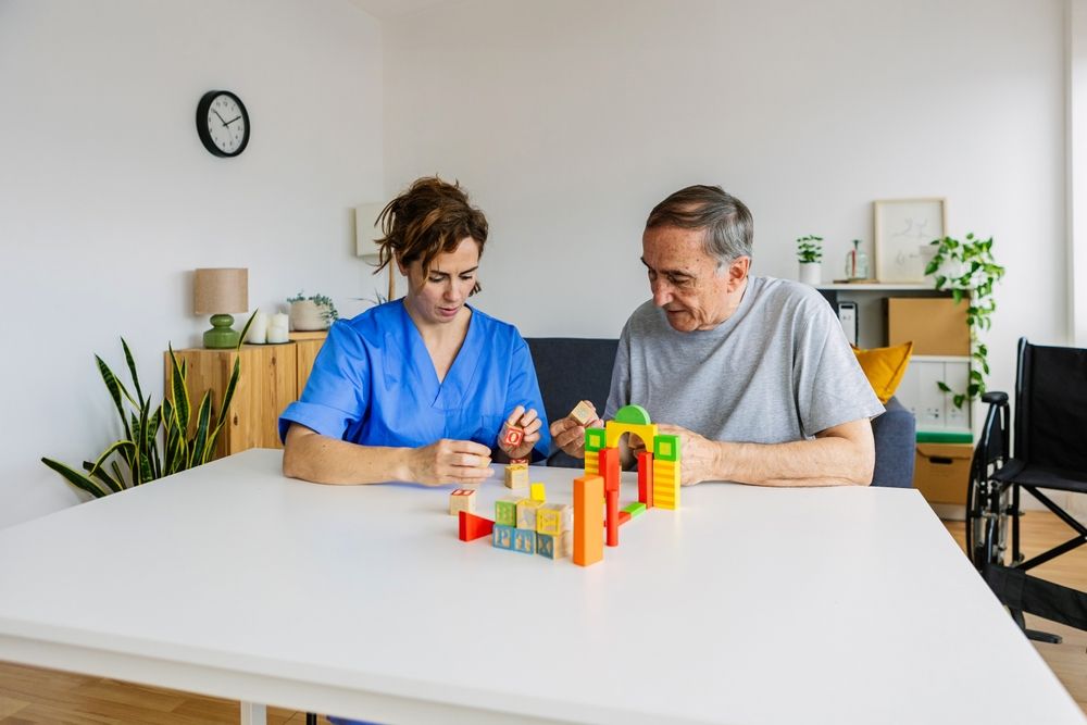Therapist in blue scrubs assists a patient building with colorful wooden blocks at a table indoors.