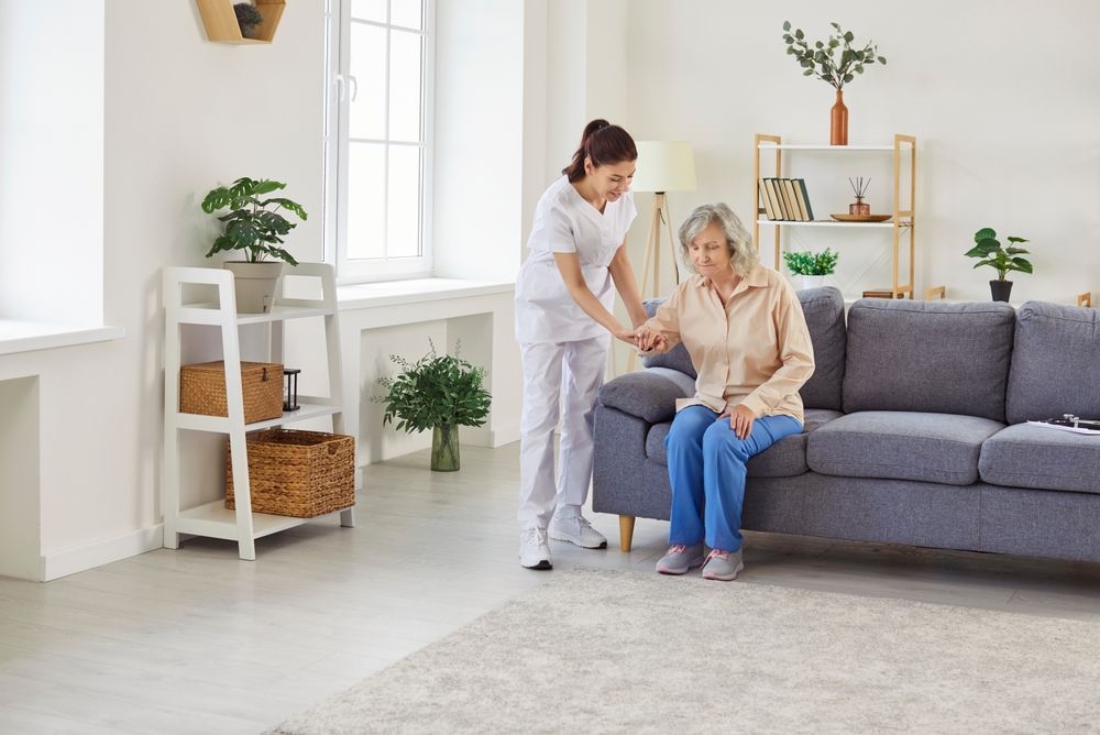 Caregiver assists person sitting on a sofa; room with light-colored walls and furniture.