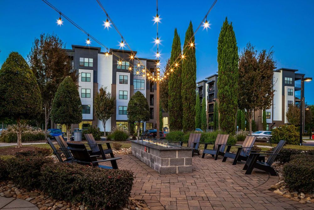 Twilight view of an outdoor seating area with chairs around a fire pit, string lights above, and modern apartment buildings in the background, surrounded by landscaped trees and shrubs at Marq at Crabtree in Raleigh, NC.