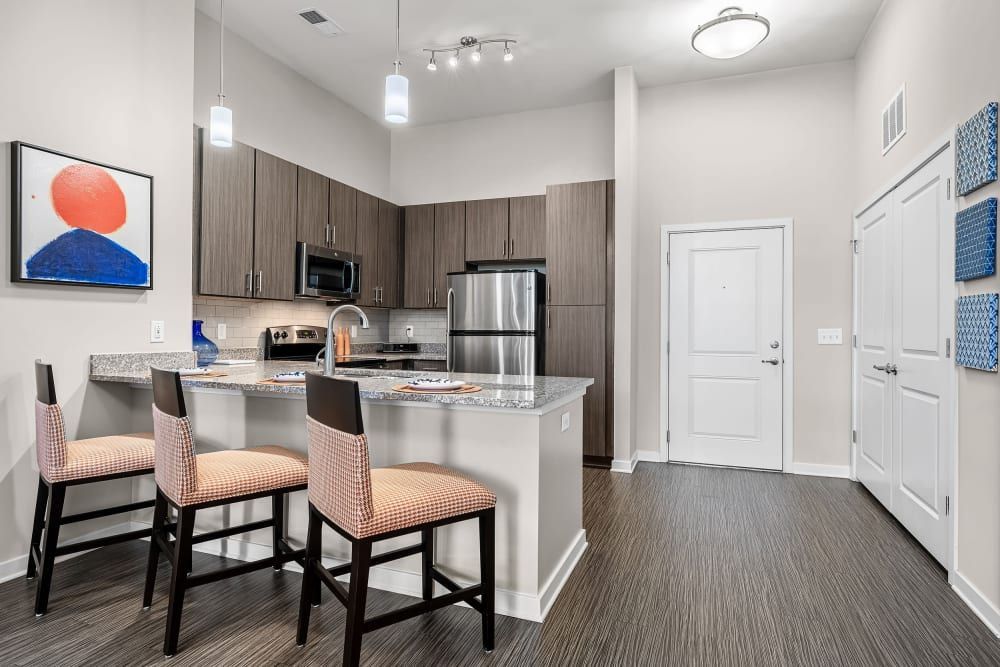 Modern kitchen interior with dark wood cabinets, stainless steel appliances, and a breakfast bar with two high chairs. The room features light walls, a decorative abstract painting, blue kitchen accessories, and a warm wooden floor at Marq at Crabtree in Raleigh, NC.