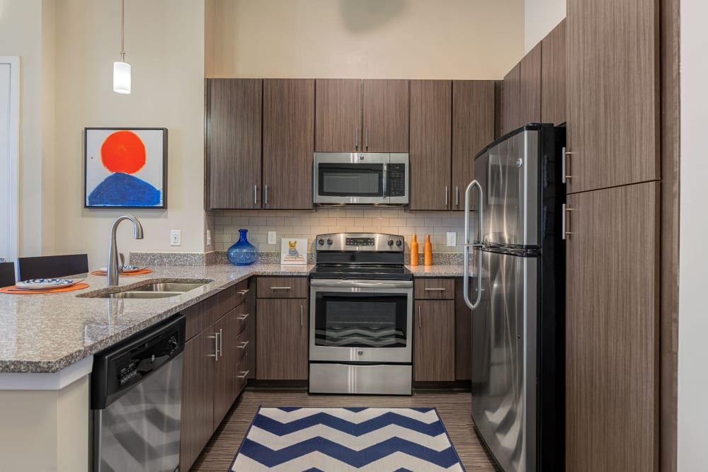 Modern kitchen interior with dark wood cabinetry, stainless steel appliances, and granite countertops. A bright abstract painting hangs on the wall, and decorative items such as a blue vase and orange dish soap add pops of color. A chevron-patterned rug lays on the floor at Marq at Crabtree in Raleigh, NC.