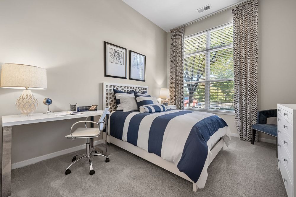A modern and neatly arranged bedroom featuring a white bed with blue and white striped bedding, and a variety of pillows. A desk with a white chair, table lamp, and decorative items is placed by the window, which is dressed with patterned curtains. Artwork hangs on the wall above the bed, and a cozy bench sits at the foot of the bed, while a small globe and picture frame adorn the desk area. The space is well-lit with natural light streaming through the window, complementing the soft grey carpet and neutral wall color at Marq at Crabtree in Raleigh, NC.