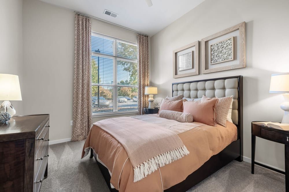 A well-lit and inviting bedroom features a large window with blinds and patterned drapes. The room is furnished with a dark wood bed frame topped with an upholstered headboard, dressed with a peach-colored bedding set and complemented by matching decorative pillows. Beside the bed are matching nightstands with lamps, and above the bed hangs a pair of framed decorative wall art pieces. The neutral carpet and walls create a warm and cozy atmosphere at Marq at Crabtree in Raleigh, NC.