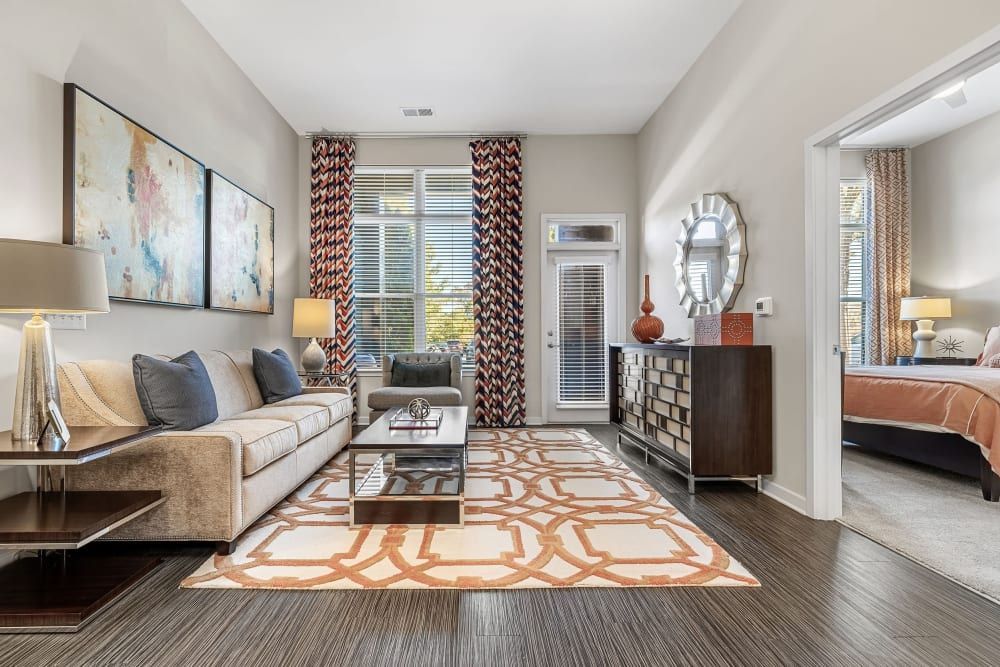 Modern living room interior with a beige upholstered sofa adorned with blue and neutral pillows, a sleek glass coffee table with a metal frame, and a large abstract painting on the wall. The room features geometric and colorful patterned curtains and a matching area rug with orange and beige tones. A decorative floor lamp, table lamp, and framed mirror add accents to the space. In the background, there is a glimpse into a bedroom with a coordinating color scheme and decor. The room is well-lit, with natural light filtering through the blinds of the windows at Marq at Crabtree in Raleigh, NC.