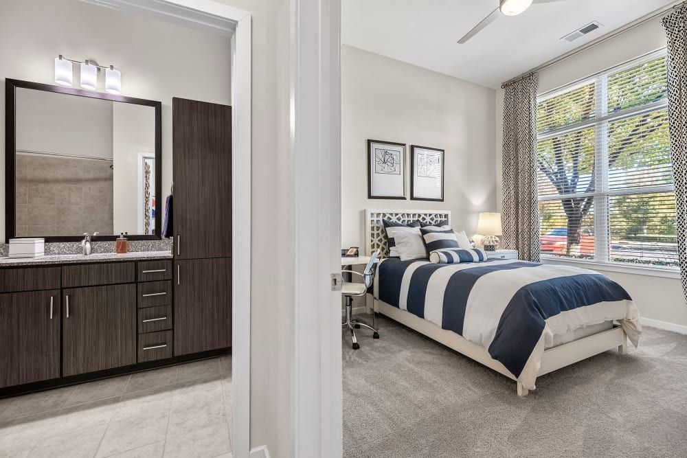 Modern bedroom with an en-suite bathroom viewed from a doorway. The bedroom features a bed with navy and white striped bedding, a matching accent pillow, and framed artwork above the headboard. A desk and chair sit in the corner near a window with patterned curtains, providing natural light. The en-suite bathroom includes a vanity with a dark wood finish, a large mirror, and a tiled backsplash at Marq at Crabtree in Raleigh, NC.