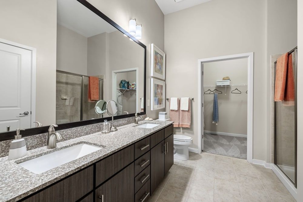 Modern bathroom interior with double vanity sink, large mirror, beige tiles, separate toilet area, and a walk-in closet. The space is well-lit, with towels and personal care items neatly arranged at Marq at Crabtree in Raleigh, NC.