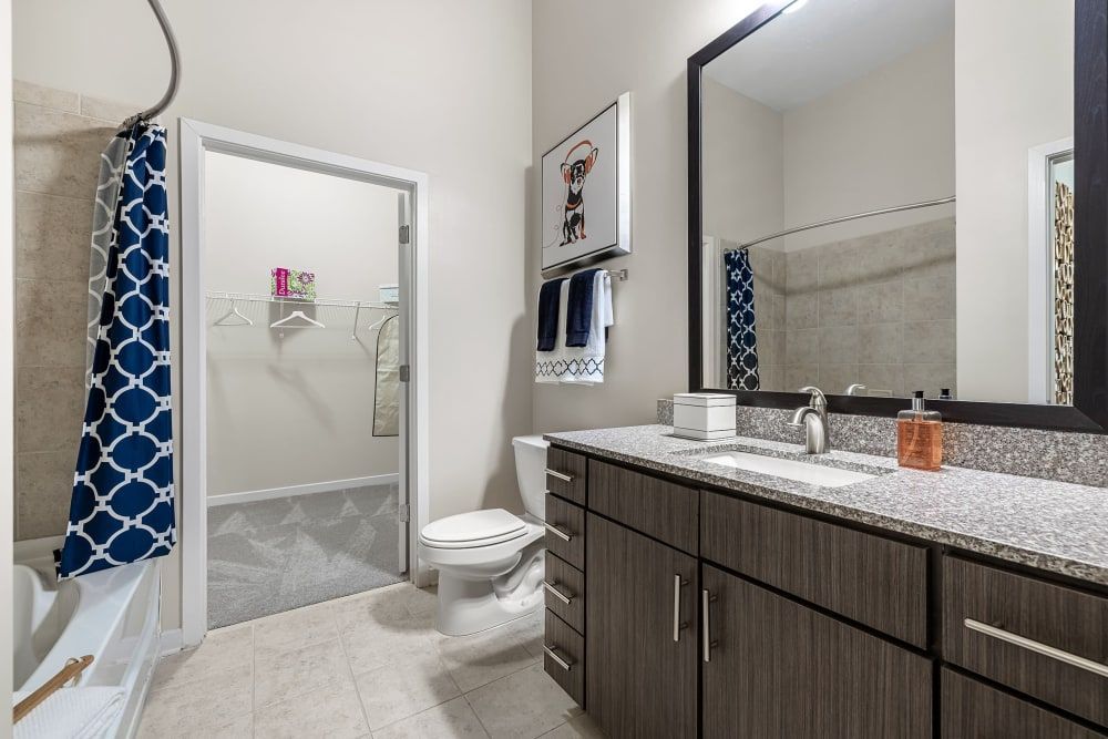 Modern bathroom with a white toilet, shower with blue patterned curtain, and a large mirror above a granite countertop with a dark wood vanity. Bathroom accessories and towels are neatly arranged, and there's artwork on the wall at Marq at Crabtree in Raleigh, NC.