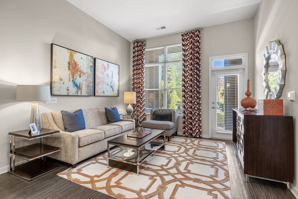 Modern living room with a beige upholstered couch adorned with blue pillows, glass-top coffee and side tables, decorative lamps, abstract wall art, and patterned curtains. The room features neutral tones with touches of blue and bronze, a wooden chest-style cabinet along one wall, a large mirror with an interesting frame design, and a door leading outside with blinds partially drawn at Marq at Crabtree in Raleigh, NC.