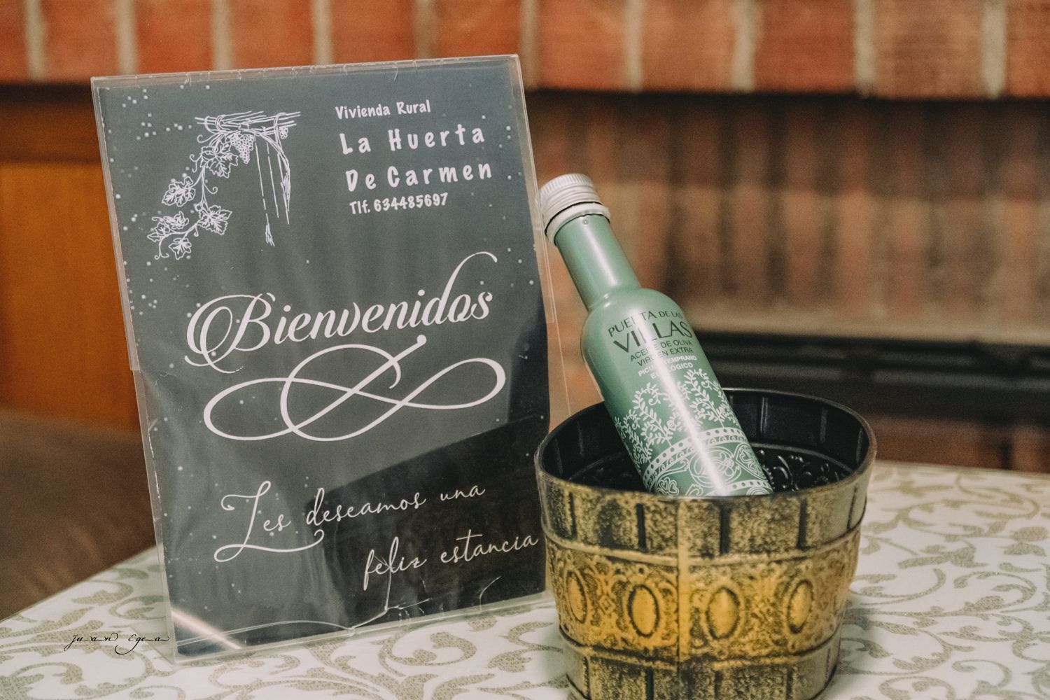 Cartel de bienvenida y botella en cubo de hielo sobre un mantel estampado, fondo de ladrillo.