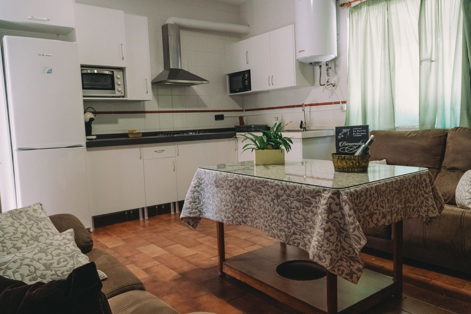 Cocina blanca con refrigerador, horno y mesa con mantel floral. Un sofá marrón cerca.