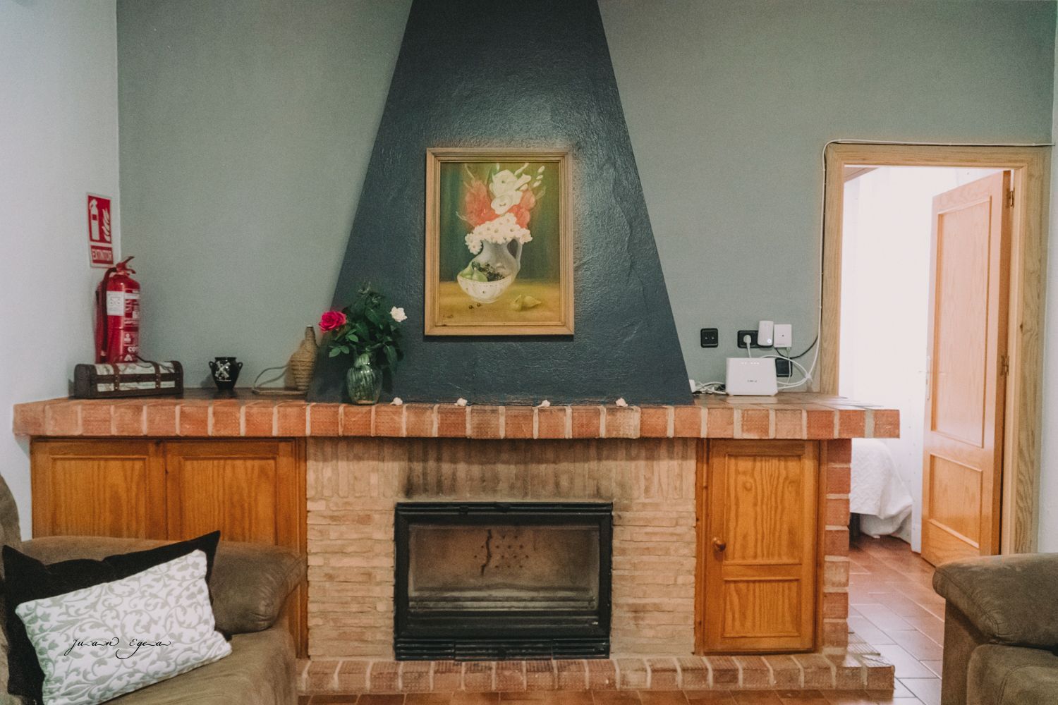 Acogedora sala de estar con chimenea de ladrillo, paredes grises y un cuadro. Una puerta da acceso a un dormitorio con dos sofás.