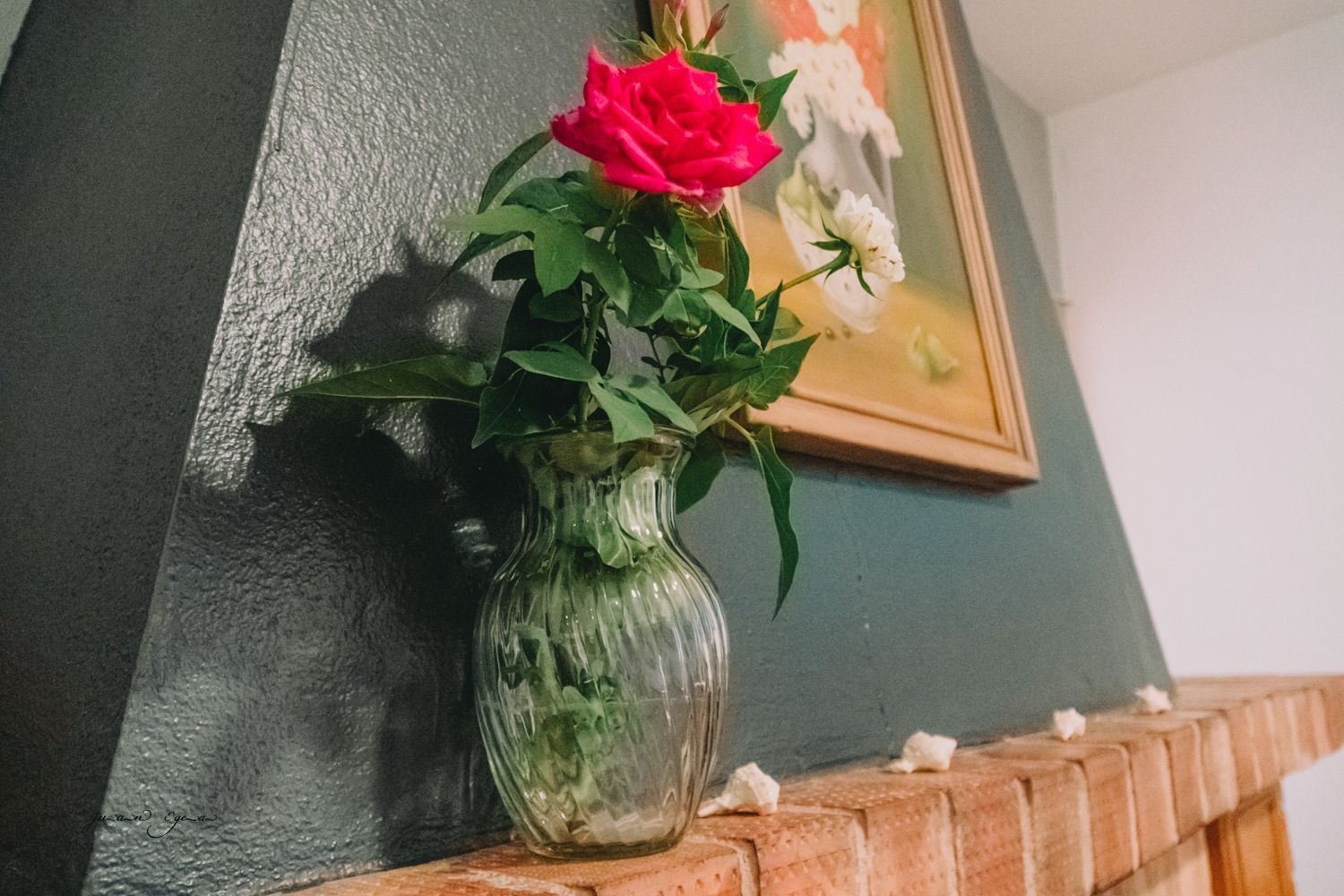 Un jarrón de cristal con rosas sobre una repisa de ladrillo. Un cuadro cuelga encima, contra una pared verde oscuro.