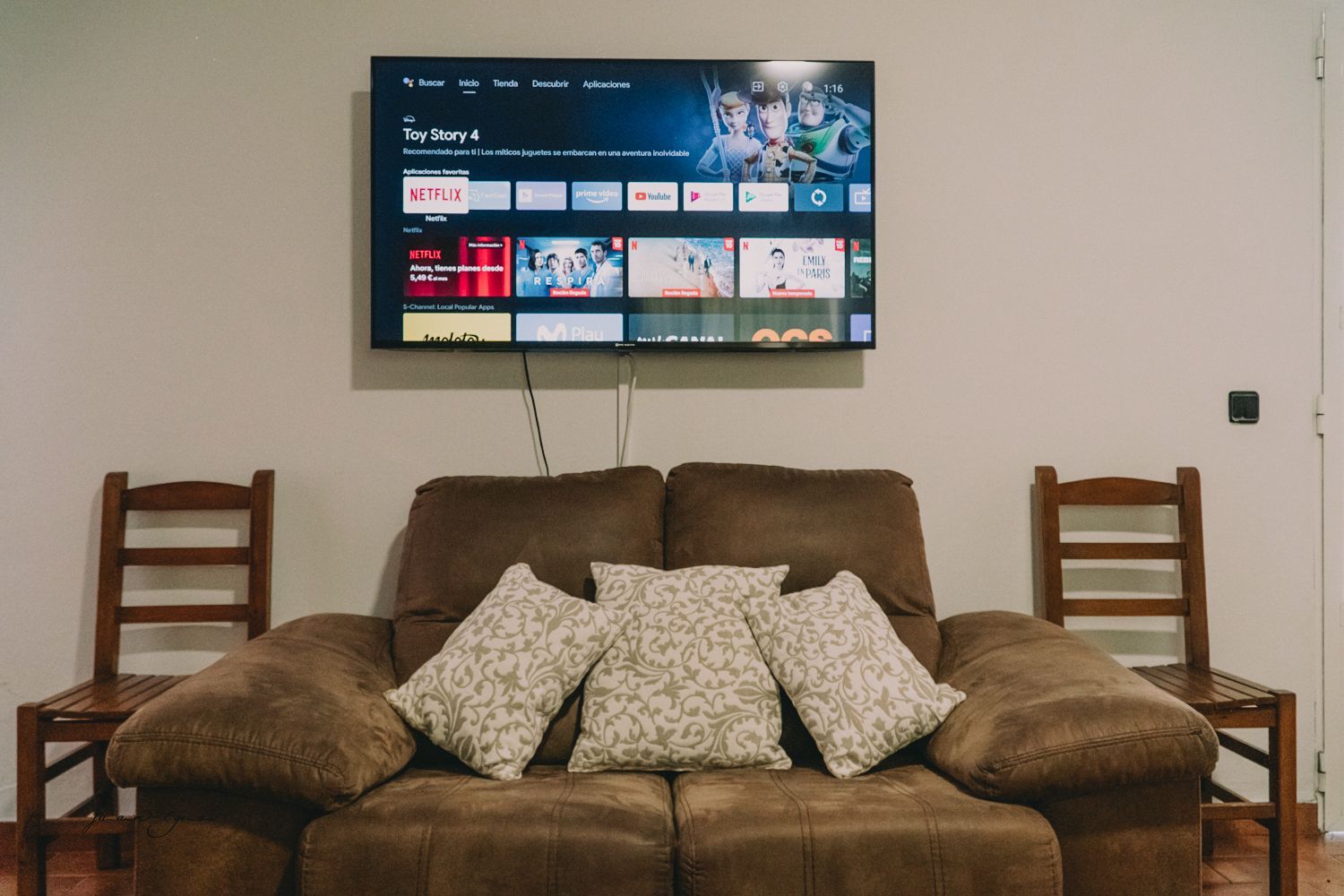 Sofá marrón, almohadas, TV en la pared, sillas de madera a ambos lados.