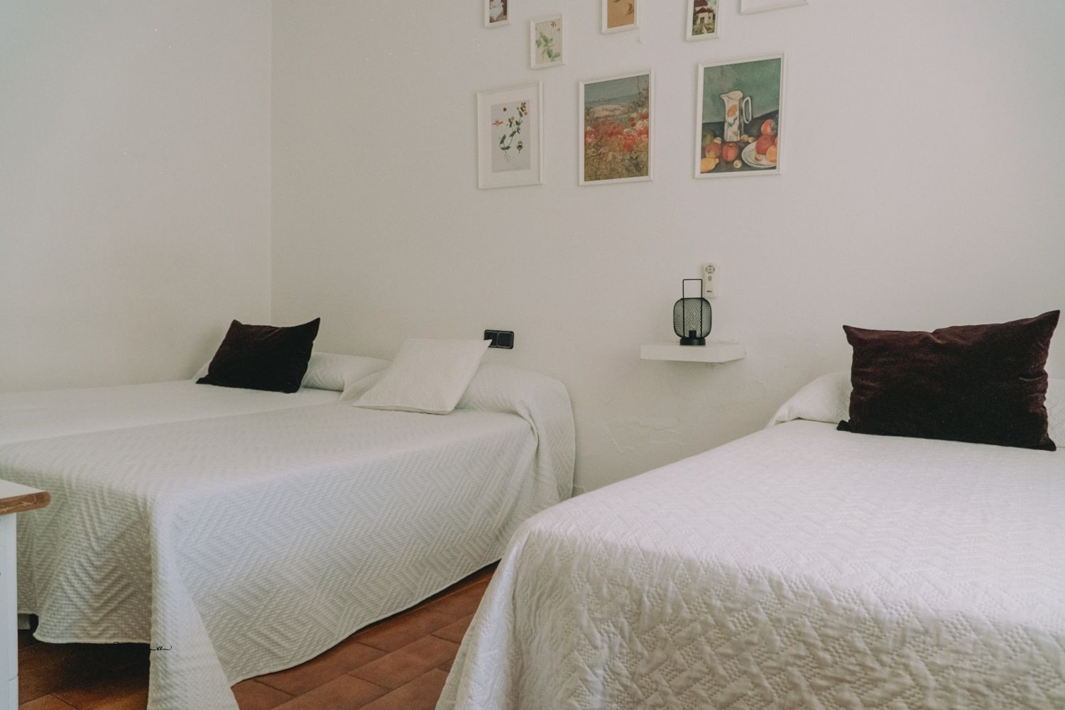 Dormitorio con dos camas individuales, ropa de cama blanca, almohadas oscuras y obras de arte enmarcadas en la pared.