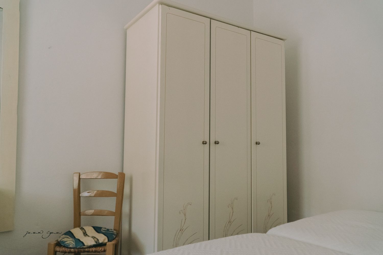 Armario color crema con tres puertas, silla y parte de cama en habitación blanca.