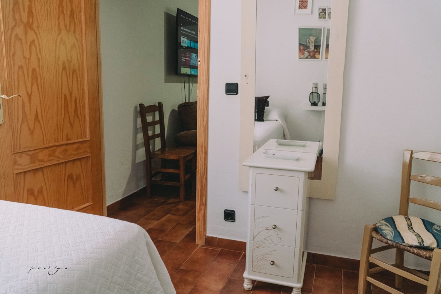 Interior de dormitorio con puerta, cómoda, espejo, televisor y sillas. Suelo de madera marrón y paredes blancas.