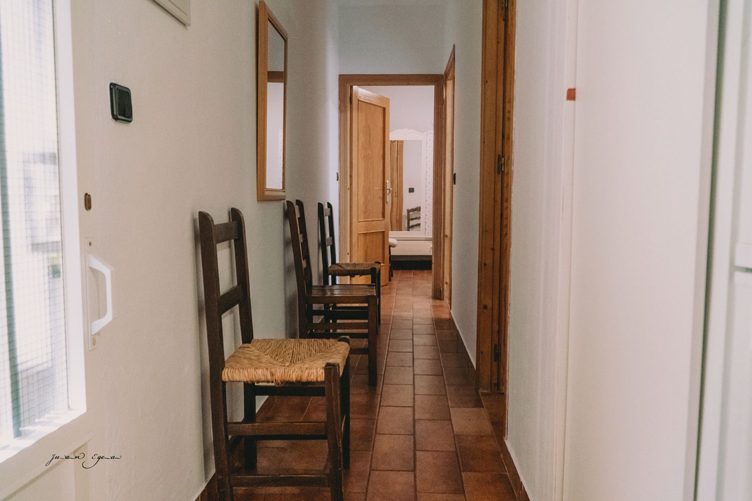 Un pasillo estrecho con tres sillas de madera. La luz entra a raudales por una ventana a la izquierda.