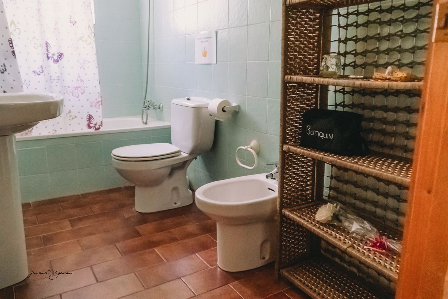 Baño con paredes turquesas, suelo de baldosas marrones y una estantería de mimbre. Inodoro, bidé y lavabo a la vista.