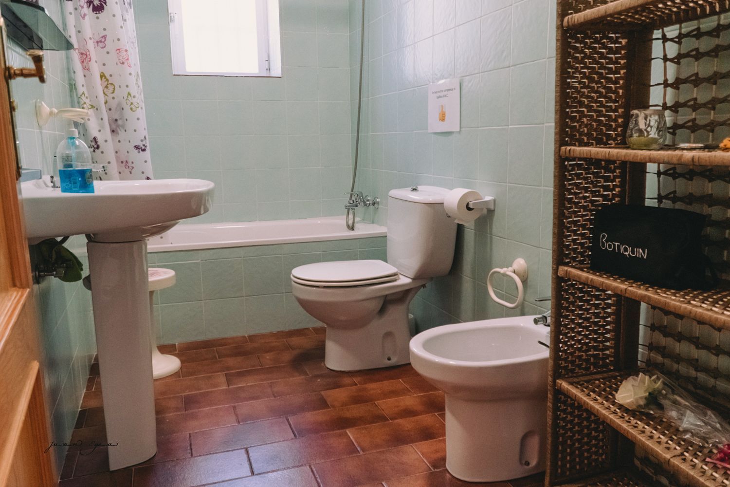 Baño con paredes alicatadas, lavabo, inodoro, bidet, bañera, estantería y suelo de madera.