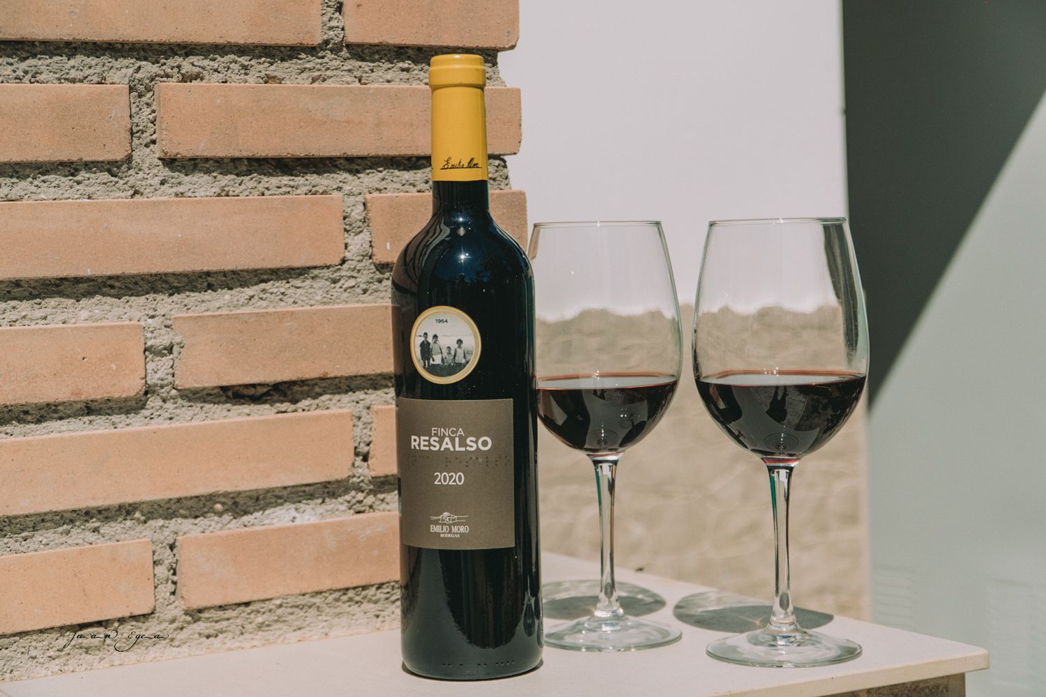 Botella de vino y dos copas llenas de vino tinto en una repisa junto a una pared de ladrillos.