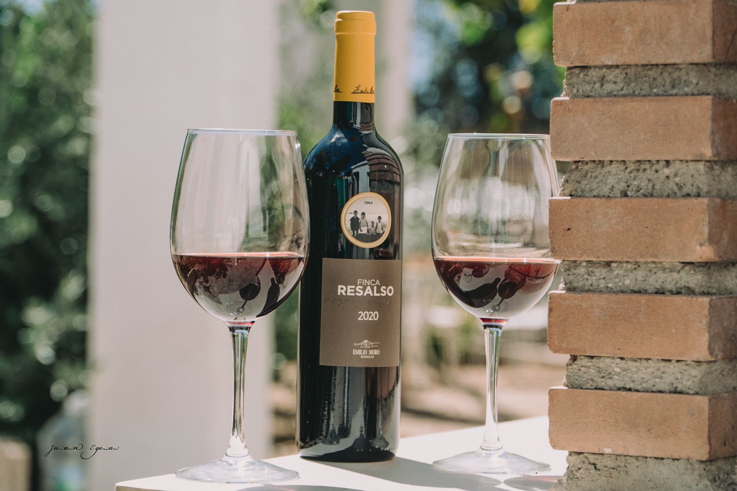 Botella de vino y dos copas llenas sobre una superficie blanca junto a una pared de ladrillos; vino tinto.