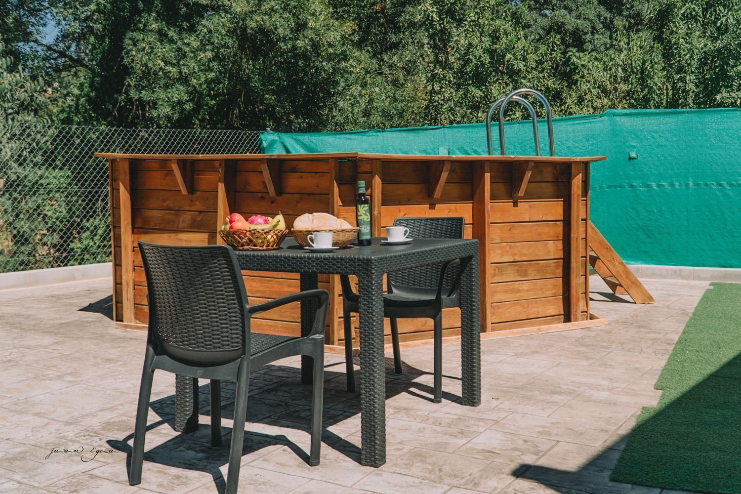 Mesa con sillas, comida y bebidas, frente a una pequeña piscina de madera, al aire libre.