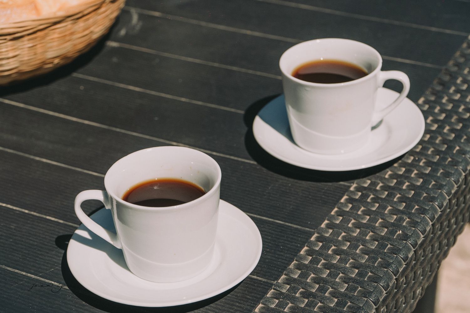 Dos tazas de café blanco llenas de café oscuro sobre una mesa negra, al lado de una canasta tejida.