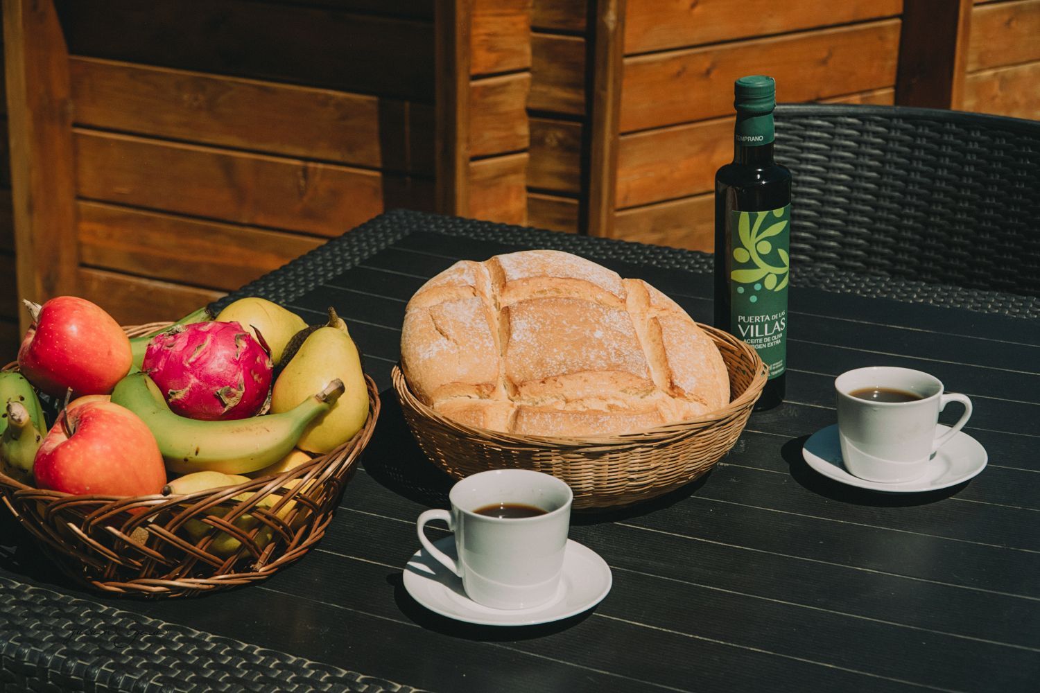 Mesa de desayuno con cesta de frutas, pan, café y botella de aceite de oliva.