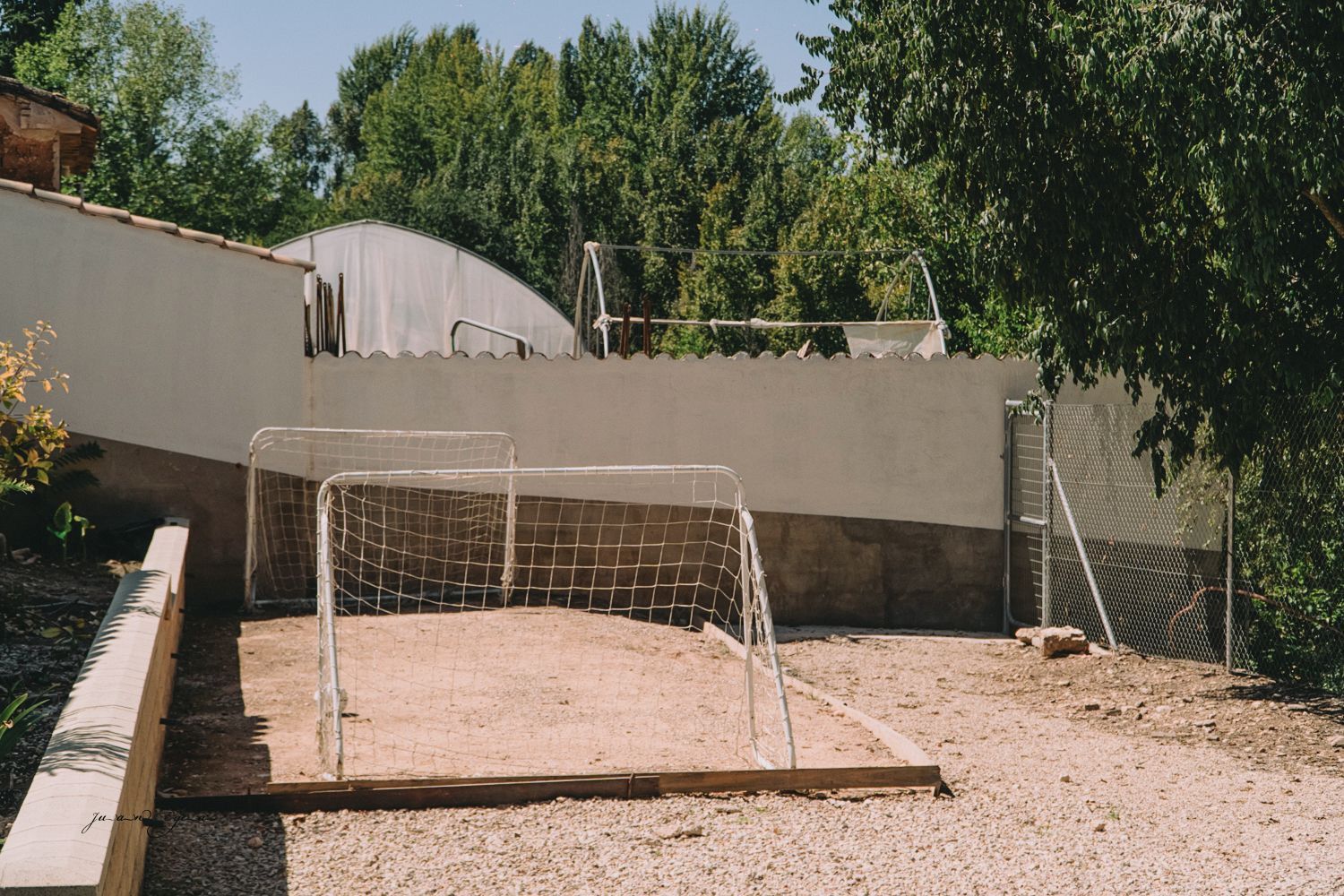 Dos pequeñas porterías de fútbol en una cancha de arena detrás de un muro, con árboles al fondo.