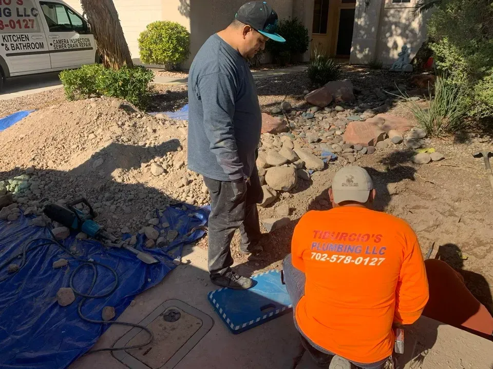 Two men work on landscaping. One kneels in an orange shirt, the other stands, looking down. Dirt, rocks, and a truck.