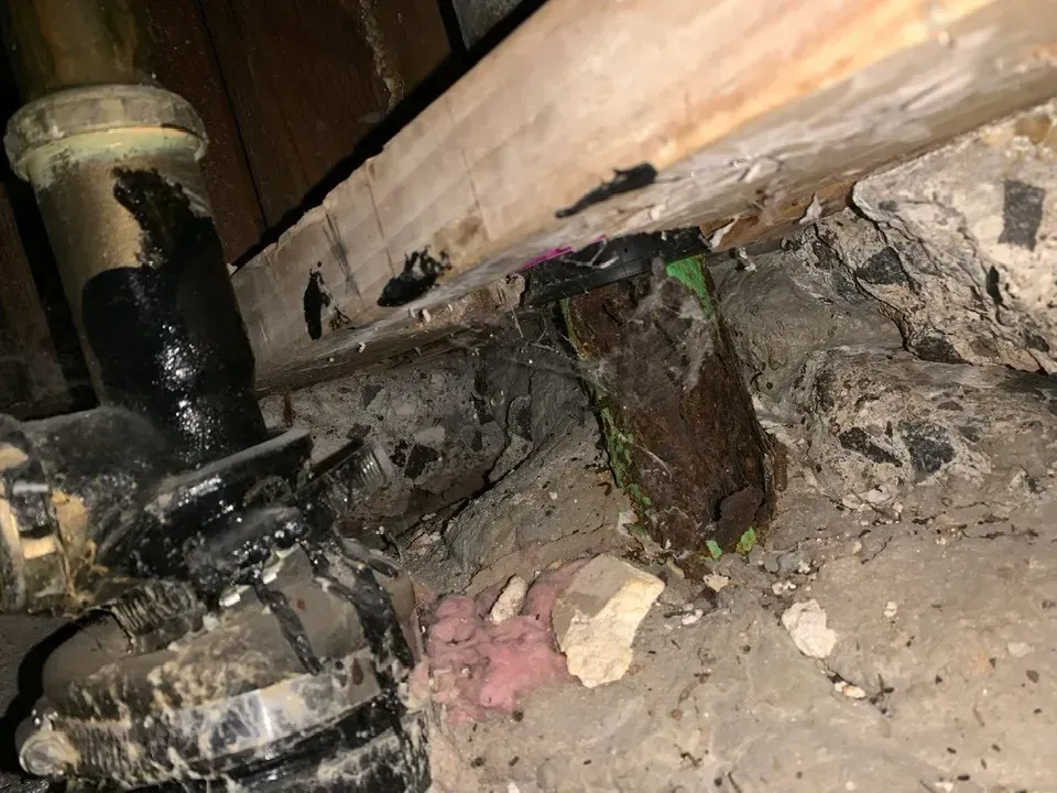Piping with black sealant leaks near damaged wood, possible water damage with debris and a dark setting.