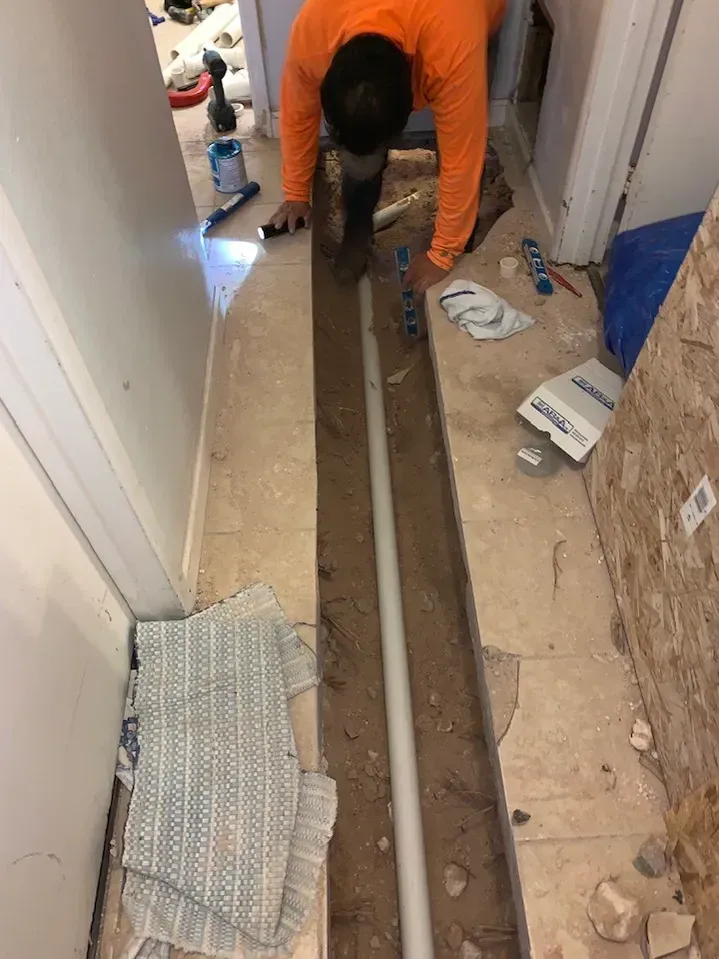Man in orange shirt working on plumbing in a trench in a hallway, with pipe and tools.