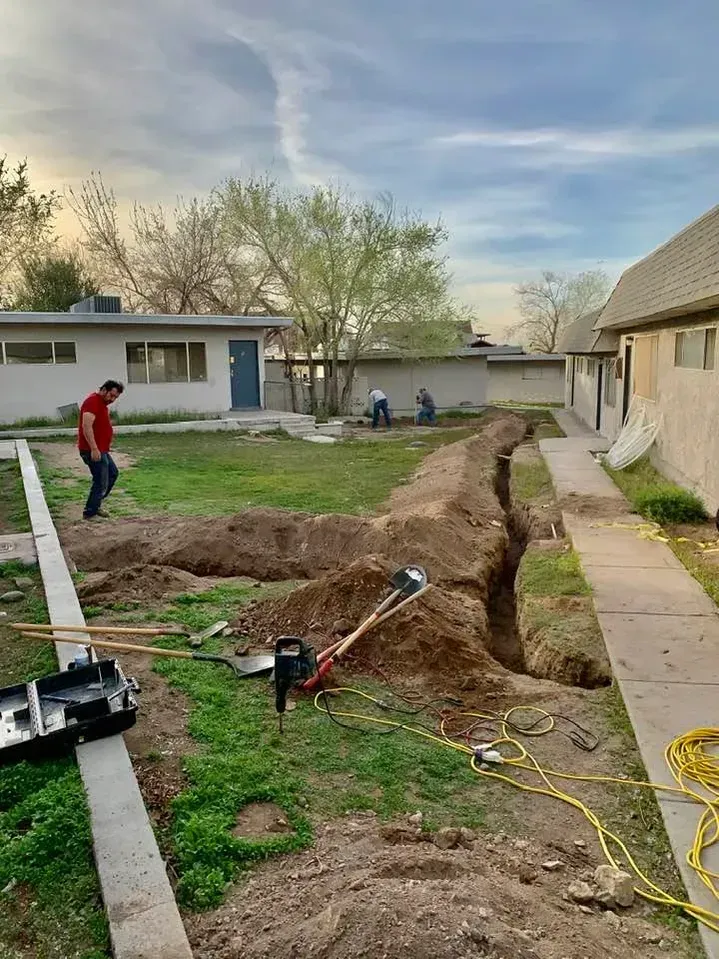 Workers dig a trench in a grassy area between buildings. One man in red shirt walks nearby.