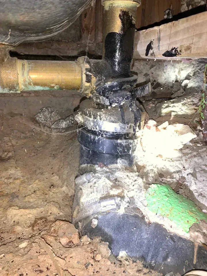 Leaking plumbing in crawlspace: black pipe with multiple clamps, brown earth, and a brass pipe.