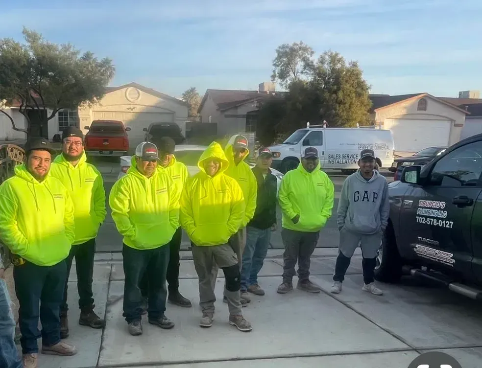 A group of men in neon hoodies stand in front of houses. A work van and truck are in the background.