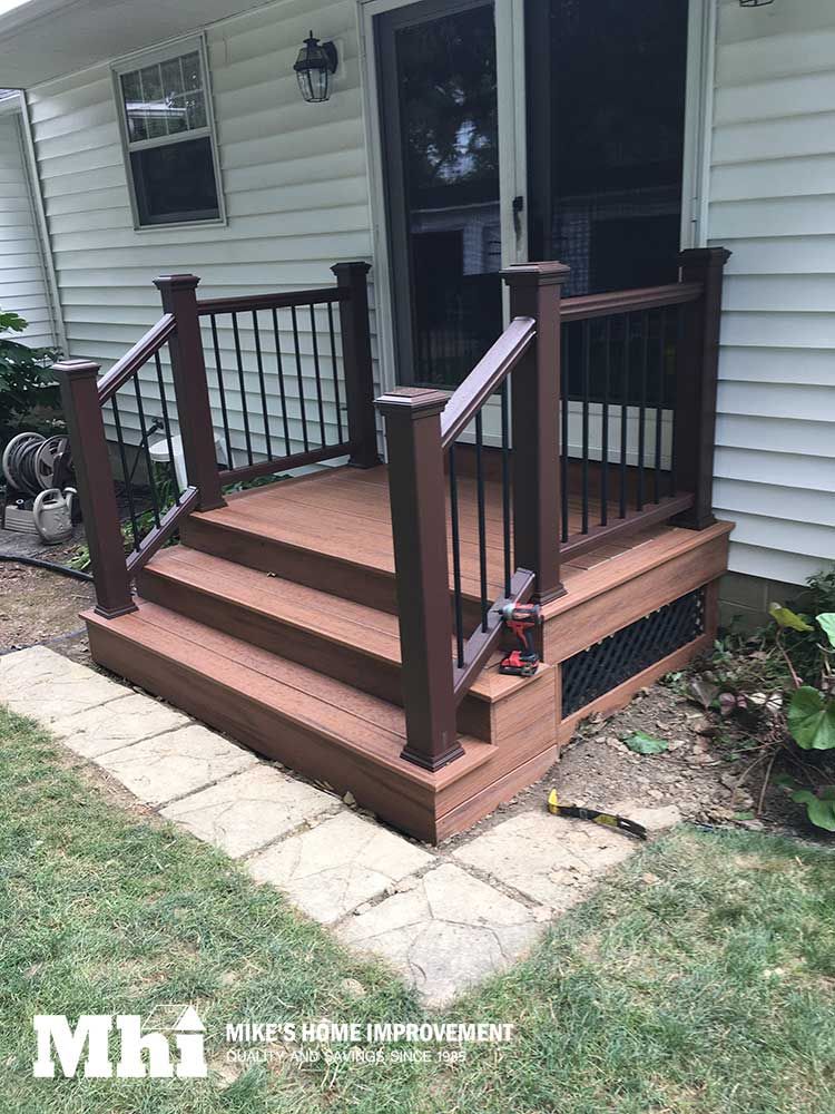 Small, newly constructed brown composite deck and stairs with black railings in front of a white house.