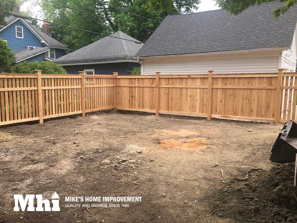 Newly installed natural wood privacy fence surrounding a bare dirt backyard with houses in the background.