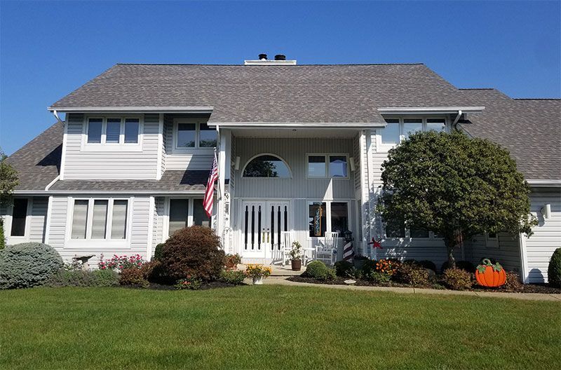 A two-story, light grey house with a dark shingled roof, front porch, and a manicured lawn under a clear blue sky.