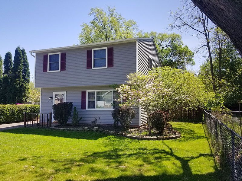A two-story house with gray siding and maroon shutters, surrounded by a lawn, trees, and a chain-link fence.