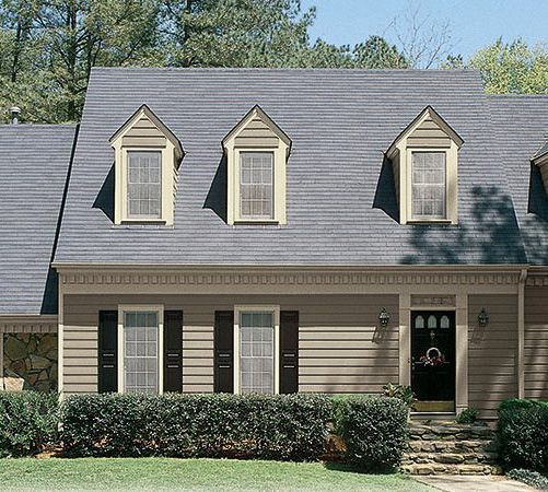 A two-story tan house with a dark gray shingled roof, three gabled dormers, black shutters, and a dark front door.
