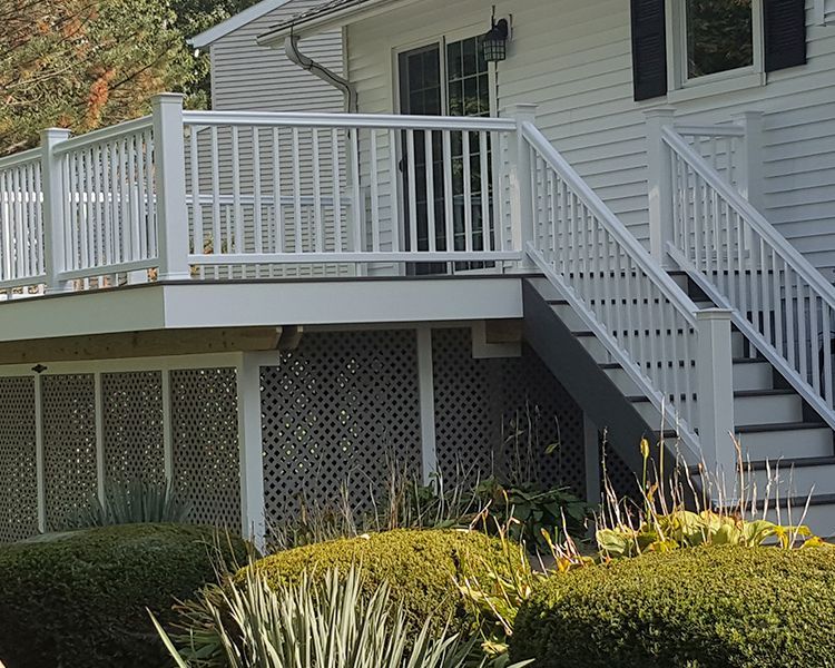 A white house features an elevated deck with white railings, stairs, and lattice skirting, surrounded by green shrubs.