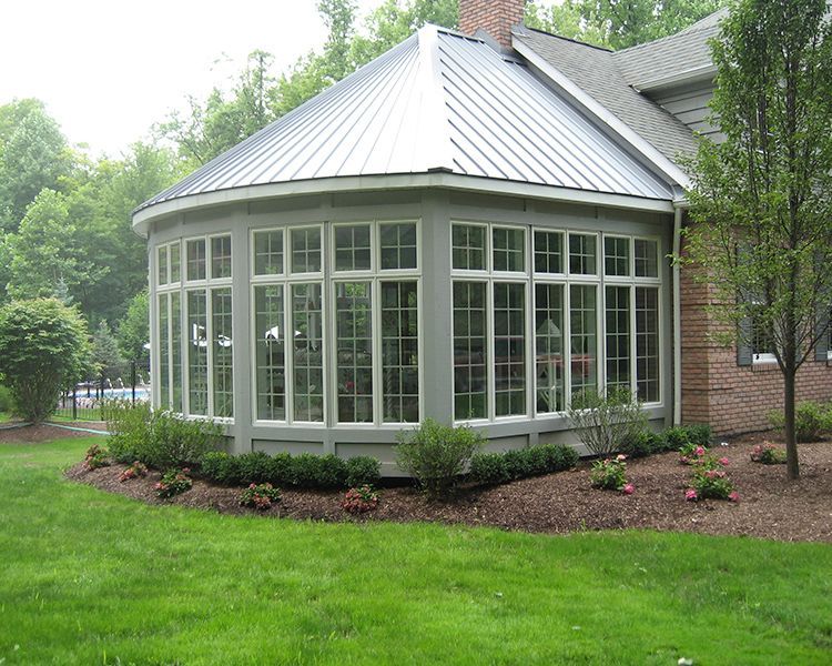 A light gray sunroom with a metal roof and large glass windows, surrounded by a lawn and landscaped garden.
