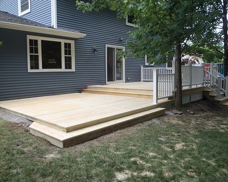 A new, multi-level light wood deck with white railings attached to the back of a blue-sided house with a large tree.